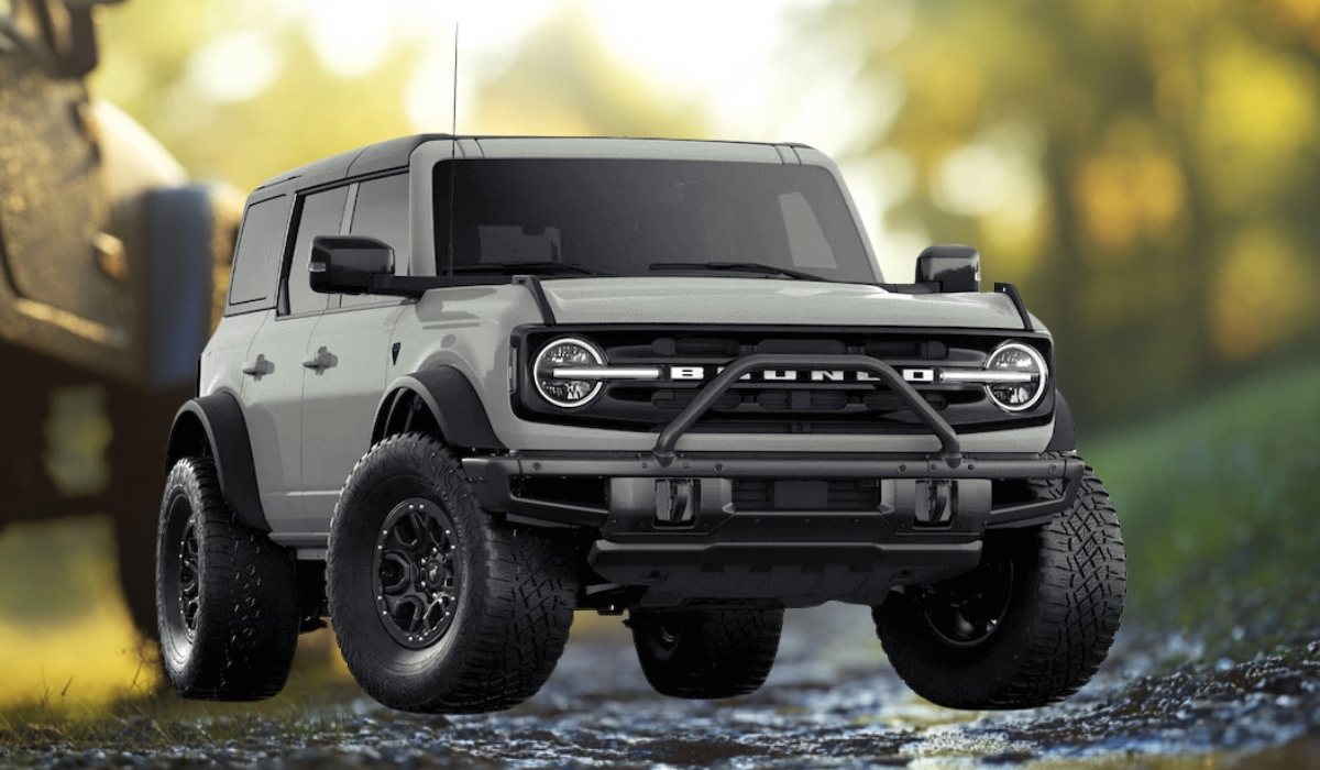 Front-facing view of a gray Ford Bronco SUV with off-road tires, parked on a forest trail with blurred trees in the background.